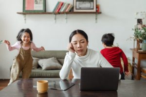 Woman at table in front of compute being stressed while two young children run around behind her